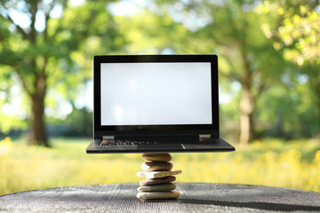 Laptop with blank screen outdoors balancing on stones surrounded by trees. Nature vs work life balance concept.