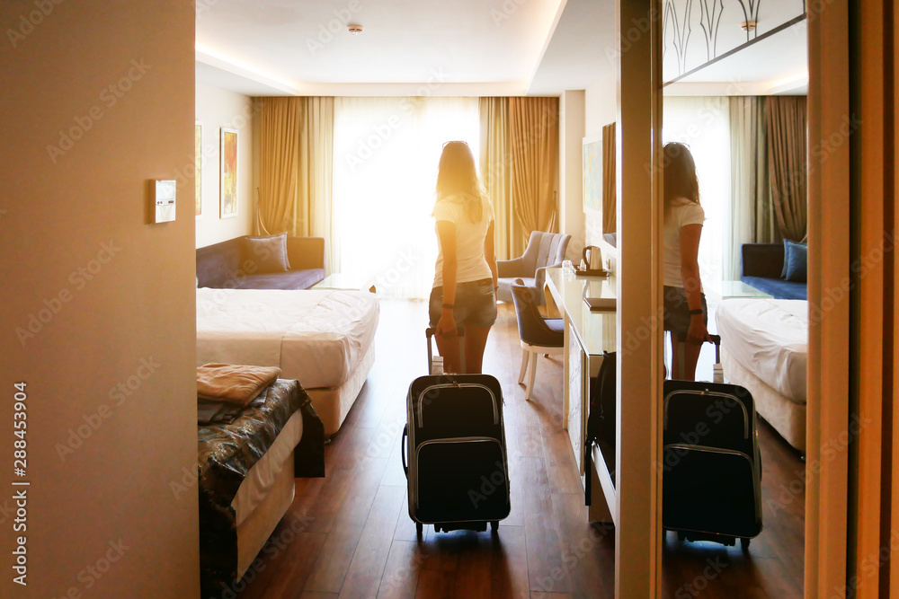 A girl comes into a bright hotel room with a suitcase on wheels. Check in at the hotel. - obrazy, fototapety, plakaty 