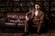young successful man in a brown kosmtyum drinks whiskey. bearded businessman sitting in a library on a luxurious leather sofa and drinking cognac