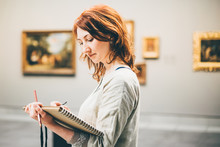 Young Woman Sketching In Museum. Blurred Pictures At The Backround.