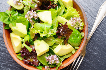 Wall Mural - Top view at clay dish with salad of avocado, green and violet lettuce, lamb's lettuce and oregano flowers on slate stone tray with fork aside