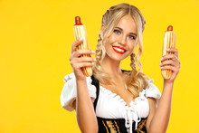 Young Sexy Oktoberfest Girl Waitress, Wearing A Traditional Bavarian Or German Dirndl With French Hot Dogs Isolated On Yellow Background.