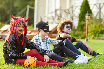 Wall Mural - Full length portrait multi-ethnic group of kids wearing Halloween costumes sitting on green lawn outdoors, copy space