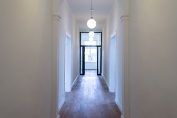 Wall Mural - A large hall in a high room with white walls, three chandeliers visible and an open glass door at the end of the corridor illuminated by the light from the window.