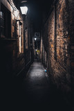 Fototapeta Uliczki - A dark creepy narrow European alley at night, surrounded by bricks and cobblestone. Illuminated only with some street lamps. Concept of scared or being alone and frightened