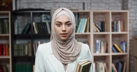 Wall Mural - Female muslim student in library, holding books and looking at camera, girl wearing hijab spending time studying - modern islam, student lifestyle concept close up 4k