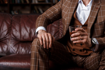 young successful man in a brown kosmtyum drinks whiskey. bearded businessman sitting in a library on a luxurious leather sofa and drinking cognac