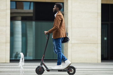 Side view full length of modern African-American businessman riding electric scooter while commuting to work in city, copy space