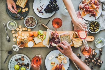 Wall Mural - Mid-summer picnic with wine and snacks. Flat-lay of charcuterie and cheese board, rose wine, nuts, olives and peoples hands holding food and celebrating over concrete table background, top view
