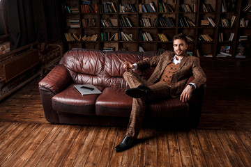 young successful man in a brown kosmtyum drinks whiskey. bearded businessman sitting in a library on a luxurious leather sofa and drinking cognac