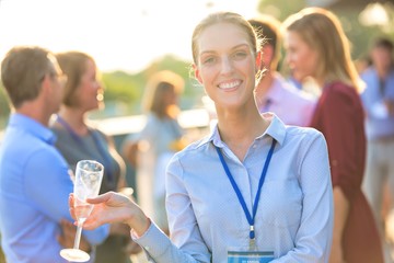 Wall Mural - Portrait of young smiling businesswoman standing while holding wine glass against colleagues at success party on rooftop