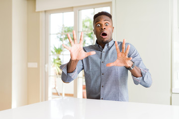 Poster - Handsome african american man on white table afraid and terrified with fear expression stop gesture with hands, shouting in shock. Panic concept.