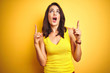© Krakenimages.com - Young beautiful woman wearing t-shirt standing over yellow isolated background amazed and surprised looking up and pointing with fingers and raised arms.