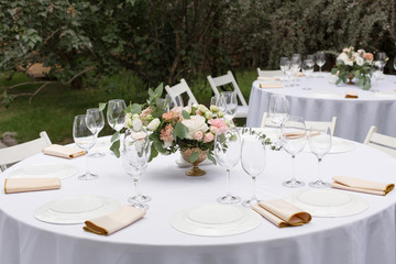 Wedding table setting decorated with fresh flowers in a brass vase. Wedding floristry. Banquet table for guests outdoors with a view of green nature. Bouquet with roses, eustoma and eucalyptus leaves