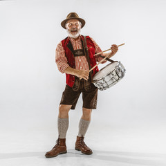 Wall Mural - Portrait of Oktoberfest senior man in hat, wearing the traditional Bavarian clothes. Male full-length shot at studio on white background. The celebration, holidays, festival concept. Playing drums.