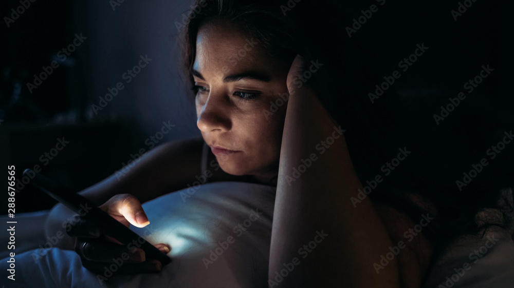 Insomnia and social media addiction concept. Young woman uses smartphone while lying in bed at night - obrazy, fototapety, plakaty 
