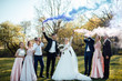 Full length portrait of newlywed couple and their friends at the wedding party with smoke checkers in green sunny park. Group of friends gathering for wedding reception. 