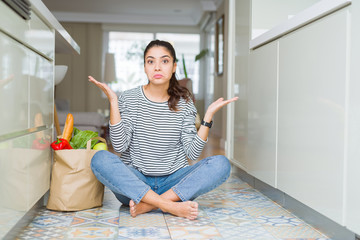 Sticker - Young woman sitting on the kitchen floor with a paper bag full of fresh groceries clueless and confused expression with arms and hands raised. Doubt concept.