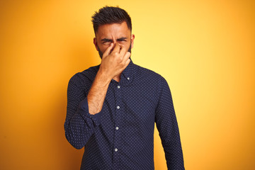 Canvas Print - Young handsome indian businessman wearing shirt over isolated yellow background smelling something stinky and disgusting, intolerable smell, holding breath with fingers on nose. Bad smells concept.