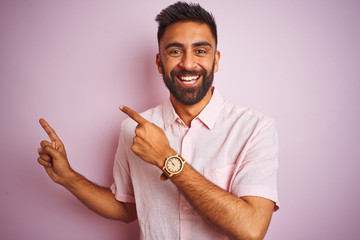 Young indian man wearing casual shirt standing over isolated pink background smiling and looking at the camera pointing with two hands and fingers to the side.