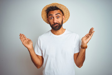 Wall Mural - Young indian man on holiday wearing summer hat standing over isolated white background clueless and confused expression with arms and hands raised. Doubt concept.