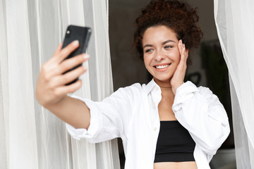 Sticker - Cheery young woman dressed in white shirt indoors in home hotel near curtain in earphones talking by mobile phone.