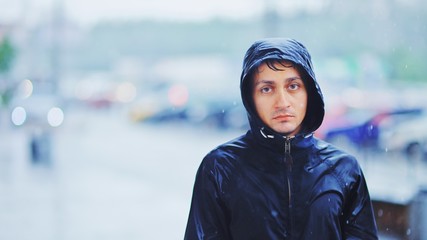 Wall Mural - Portrait of a sad man in a raincoat and a hood in the rain. Bad stormy rainy weather concept.