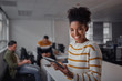 © StratfordProductions - Cheerful young african american businesswoman in casual wear holding digital tablet smiling and looking to camera while her colleagues communicating in the background - female entrepreneur - women