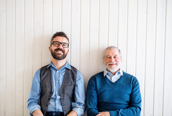 A portrait of adult hipster son and senior father sitting on floor indoors at home.