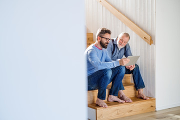 an adult son and senior father with tablet sitting on stairs indoors at home.