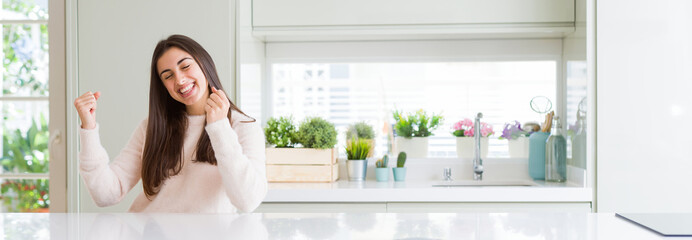 Poster - Wide angle picture of beautiful young woman sitting on white table at home very happy and excited doing winner gesture with arms raised, smiling and screaming for success. Celebration concept.