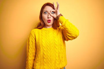 Canvas Print - Beautiful redhead woman wearing winter sweater standing over isolated yellow background doing ok gesture shocked with surprised face, eye looking through fingers. Unbelieving expression.