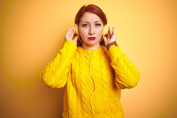 Wall Mural - Young redhead woman listening to music using headphones over yellow isolated background with serious expression on face. Simple and natural looking at the camera.