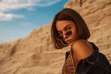 Outdoor close up portrait of young beautiful woman with tanned skin wearing snakeskin print swimwear, trendy sunglasses, hoop earrings, posing in sand desert, at sunset. Copy, empty space for text