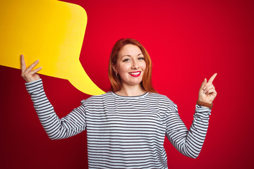 Sticker - Young beautiful redhead woman holding yellow speech bubble over red isolated background very happy pointing with hand and finger to the side