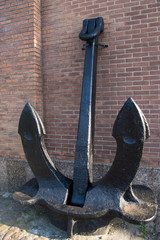 A huge vintage black metal anchor with two large pointed paws standing on the ground, propped up and attached to a red brick wall