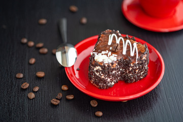 Wall Mural - A delicious heart-shaped cake sprinkled with pieces of chocolate lies on a red plate. Photographed close-up on a wooden background.