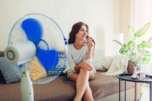 Young Woman Cooling Down By Ventilator At Home While Drinking Water And Hanging In Phone. Summer Heat.