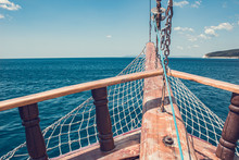 View Of The Island From The Old Wooden Ship. The Bow Of A Vintage Ship. The Wooden Stem Of A Ship, The Dangling Net, The Furled Sails And Wooden Beams Of An Ancient Ship Sailing Past The Land.