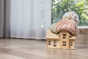 Wooden house model in hat on floor indoors, space for text. Heating efficiency