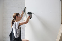 Young Woman Drilling Screws Into Plasterboard With An Electric Screwdriver, Home Improvement Concept