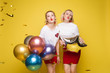 © Вячеслав Косько - Two happy beautiful woman friend celebrating posing surrounded by colorful air balloon confetti. Attractive smiling female wearing bright clothes enjoying party isolated at yellow studio background