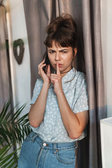 Poster - Beautiful woman talking by mobile phone indoors at home showing silence gesture.