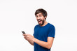 © ianachyrva - Handsome brunet man in a blue shirt with beard and mostaches surprised holding smartphone looking at the camera standing isolated over white background.
