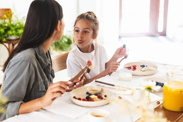 Wall Mural - Image of amusing family mother and little daughter eating tasty pancakes together while having breakfast at home in morning