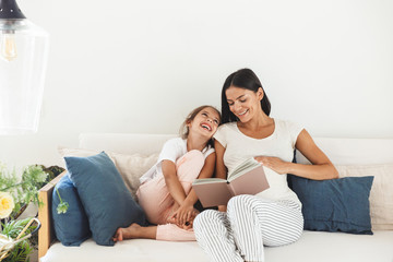 Wall Mural - image of joyful family pregnant woman and her little daughter reading book while sitting on sofa at 