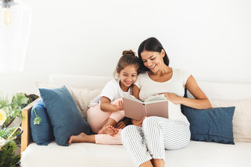 Wall Mural - image of attractive family pregnant woman and her little daughter reading book while sitting on sofa