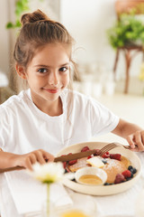 Wall Mural - Portrait of caucasian little girl sitting at table in bright kitchen while eating pancakes with raspberry