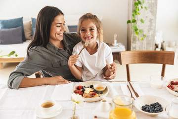 Wall Mural - image of joyous family mother and little daughter smiling and eating together while having breakfast