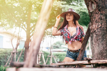 Portrait Of Caucasian Young Woman In Checkered Shirt And Cowboy Hat. She Is Looking Aside While Touching Her Hat Standing Near Tree Outdoors. Living On A Farm Concept. Horizontal Shot.